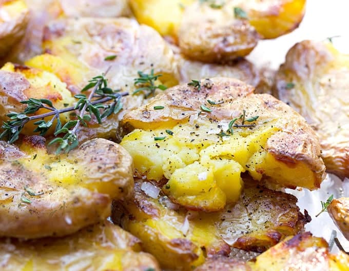 Close up of about 8 crispy smashed red-skinned, yellow-flesh potatoes, topped with chopped thyme and black pepper