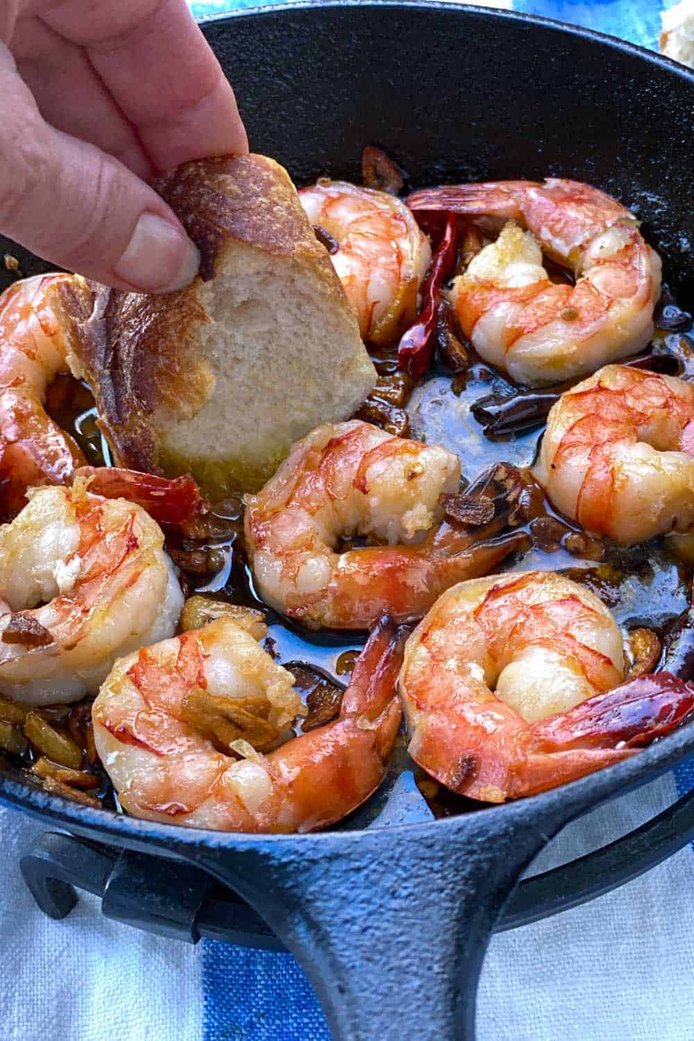 8 sauteed shrimp in a small cast iron skillet, a hand dipping a slice of baguette into the garlicky oil between the shrimp.