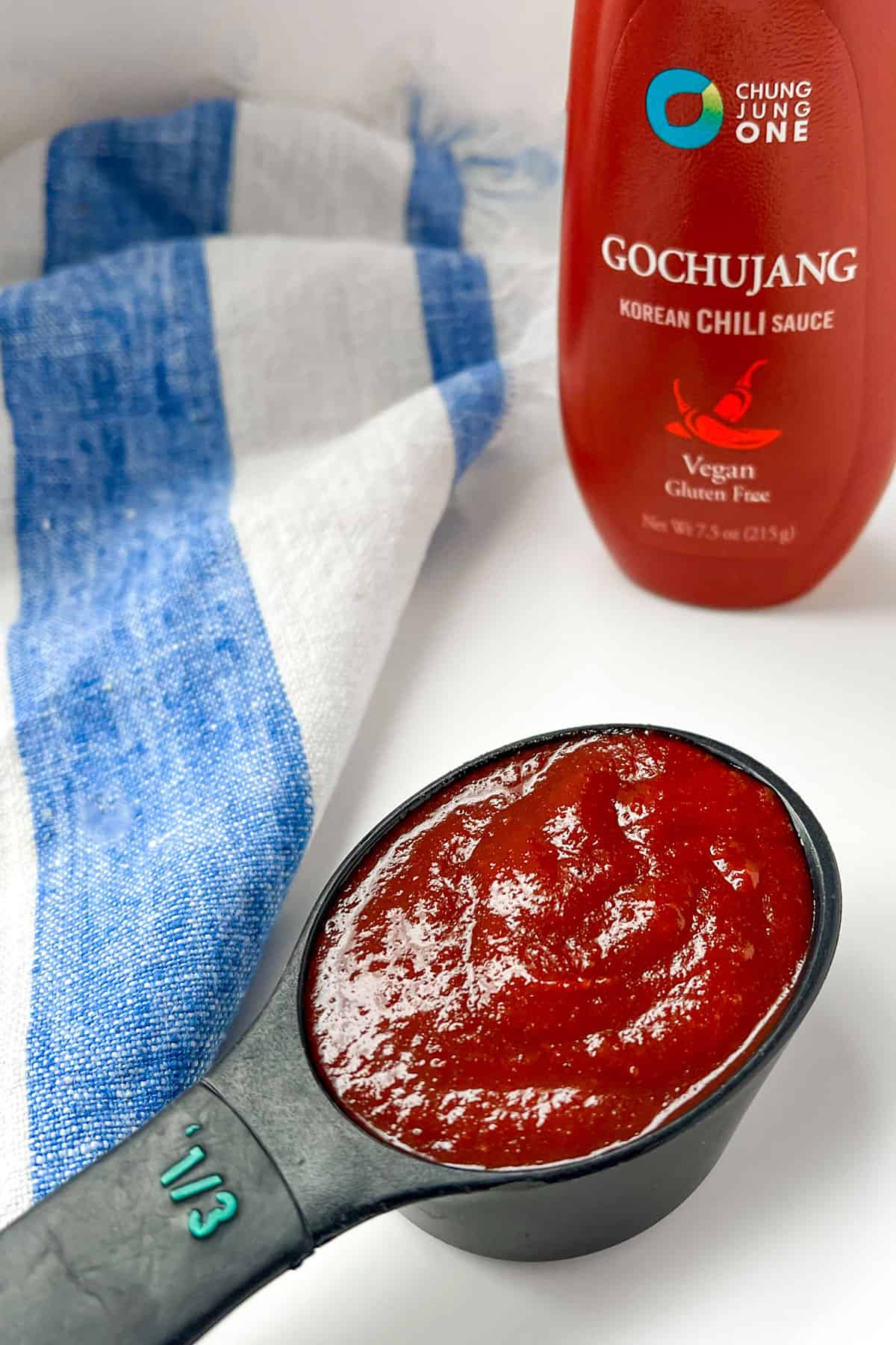 small black measuring spoon holding ⅓ cup of Korean gochujang sauce, with the bottle of the sauce in the background