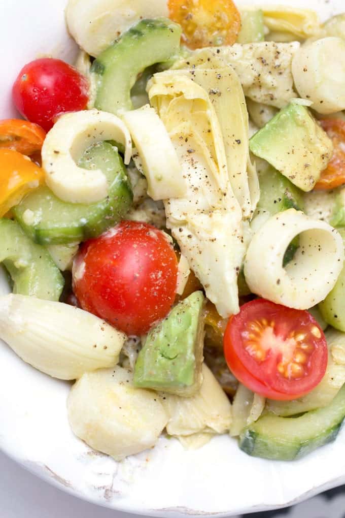 close up of a white bowl filled with sliced hearts of palm, sliced cucumbers, halved cherry tomatoes, chunks of avocado and sliced canned artichoke hearts.