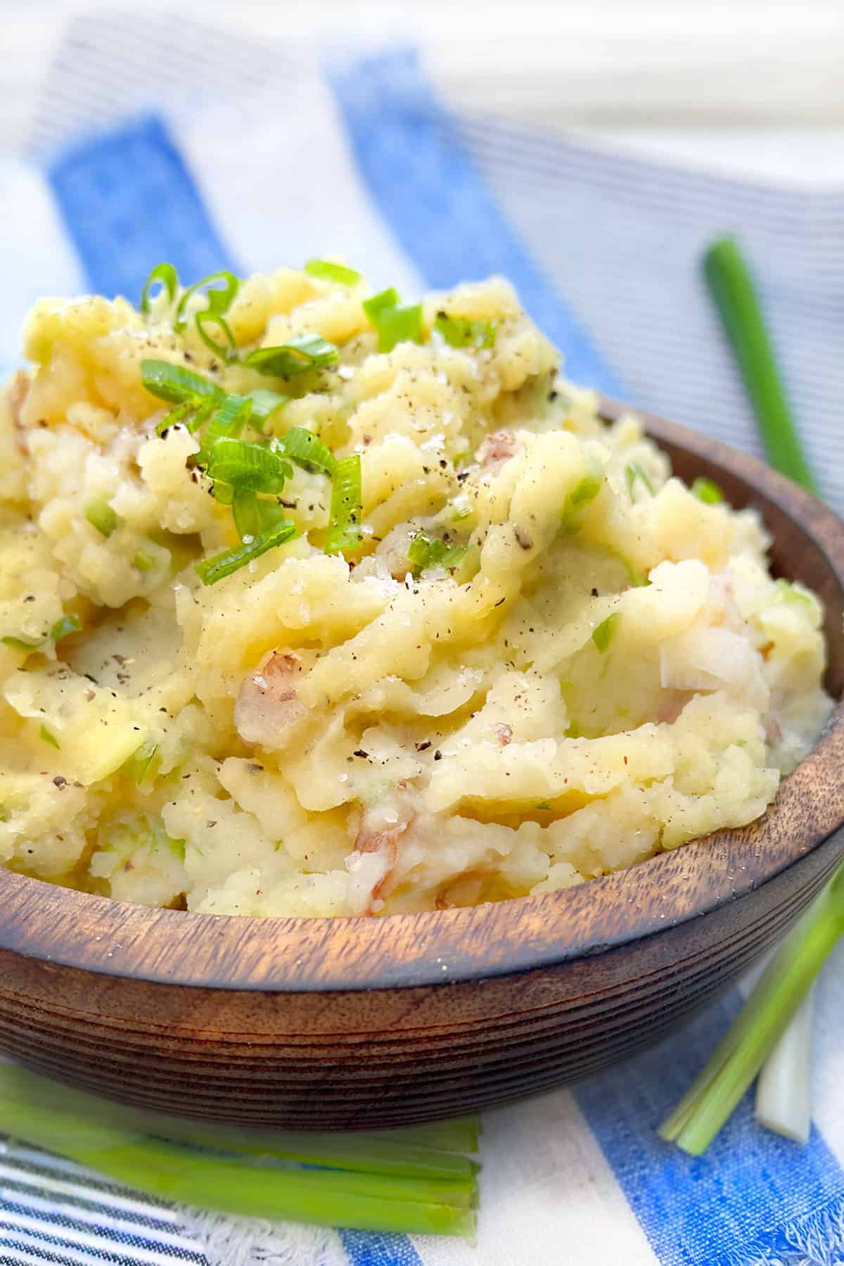mashed red skin potatoes with their skin, in a brown wooden serving bowl, topped with chopped scallions and black pepper.