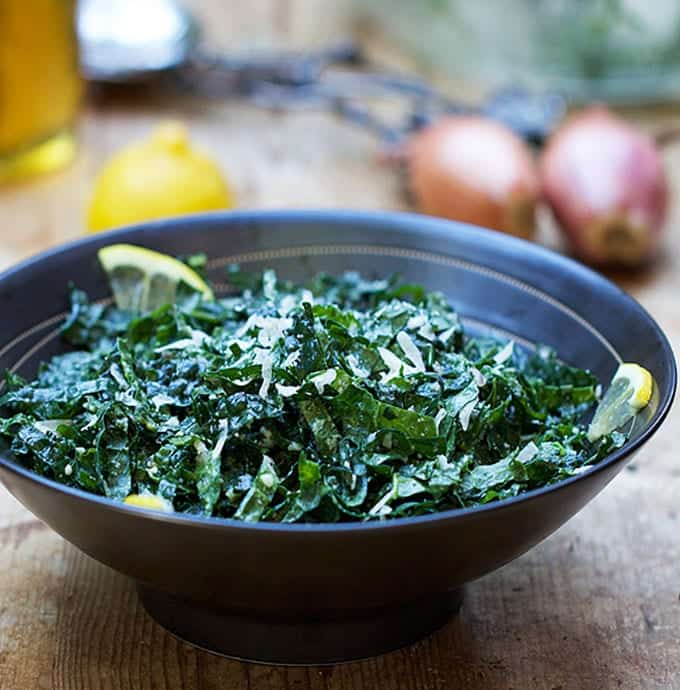 shredded kale in a black bowl topped with shreds of parmesan, lemon slices on the side of the bowl, shallots and lemons in the background