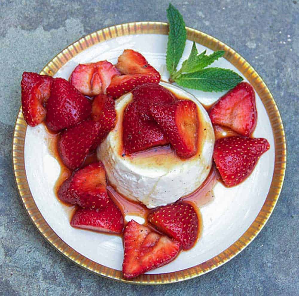 gold-rimmed white dessert plate topped with a mound of vanilla panna cotta surrounded by macerated sliced strawberries, a mint sprig for garnish.