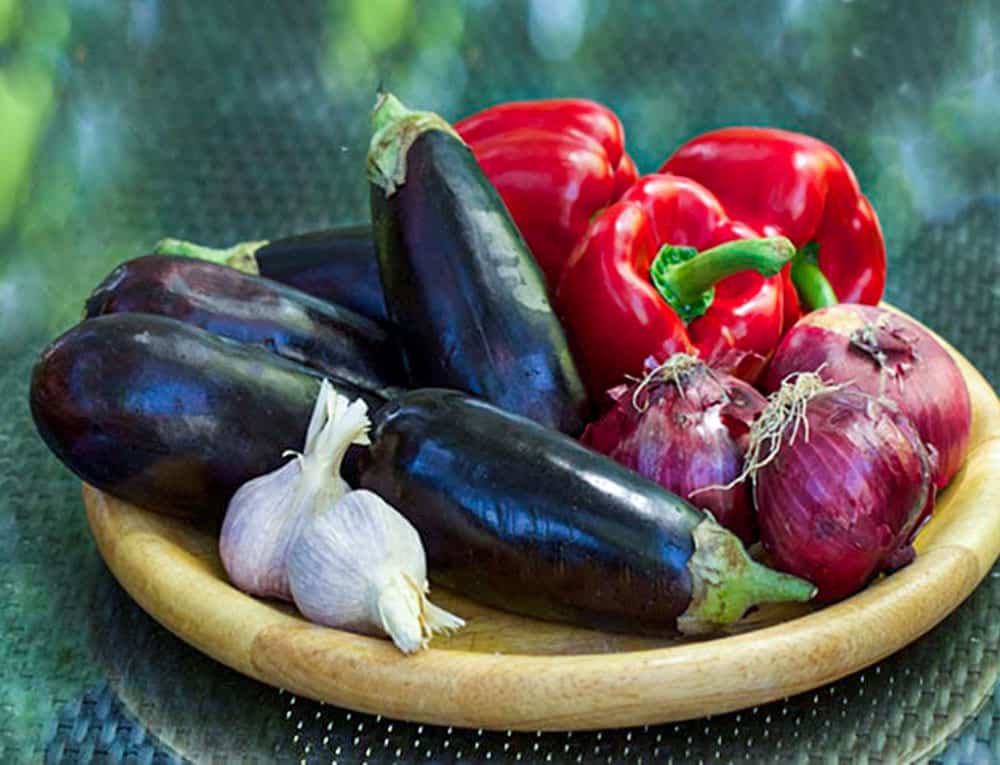 4 eggplants, 3 red bell peppers, 3 red onions, 2 heads of garlic on a wooden round tray.
