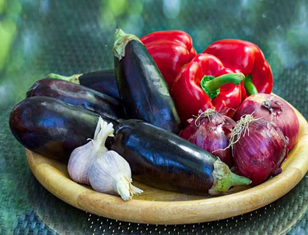 4 eggplants, 3 red bell peppers, 3 red onions, 2 heads of garlic on a wooden round tray.