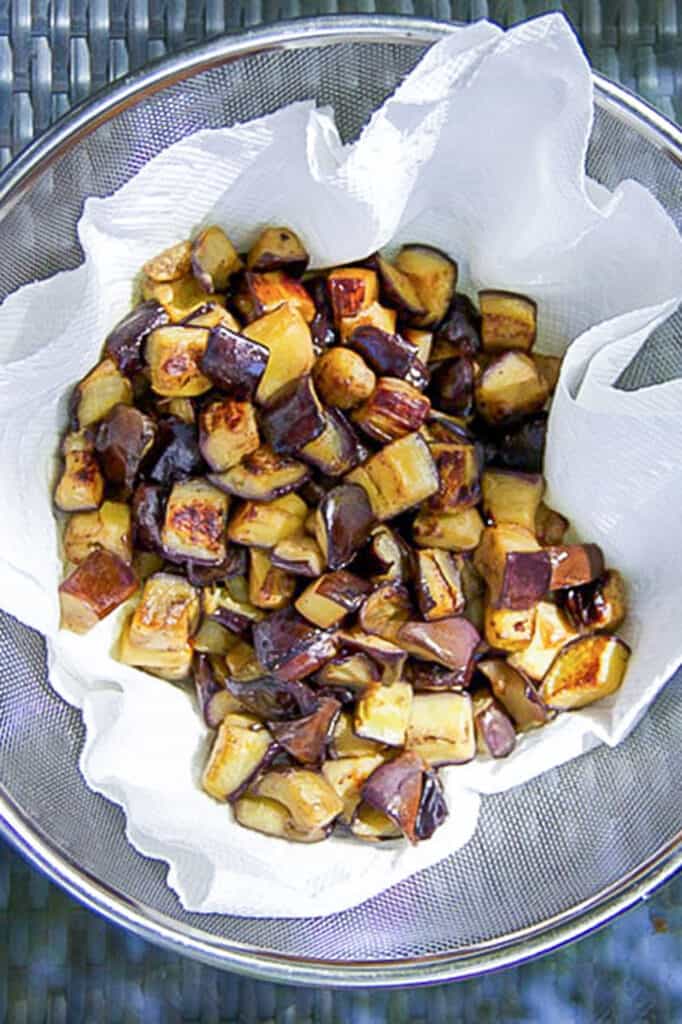browned cubes of eggplant draining in a paper towel lined mesh colander