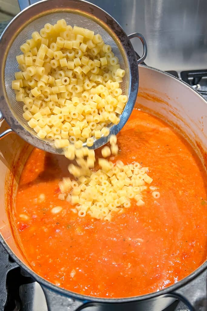 cooked Ditalini pasta being poured from a small mesh strainer into a pot of soup