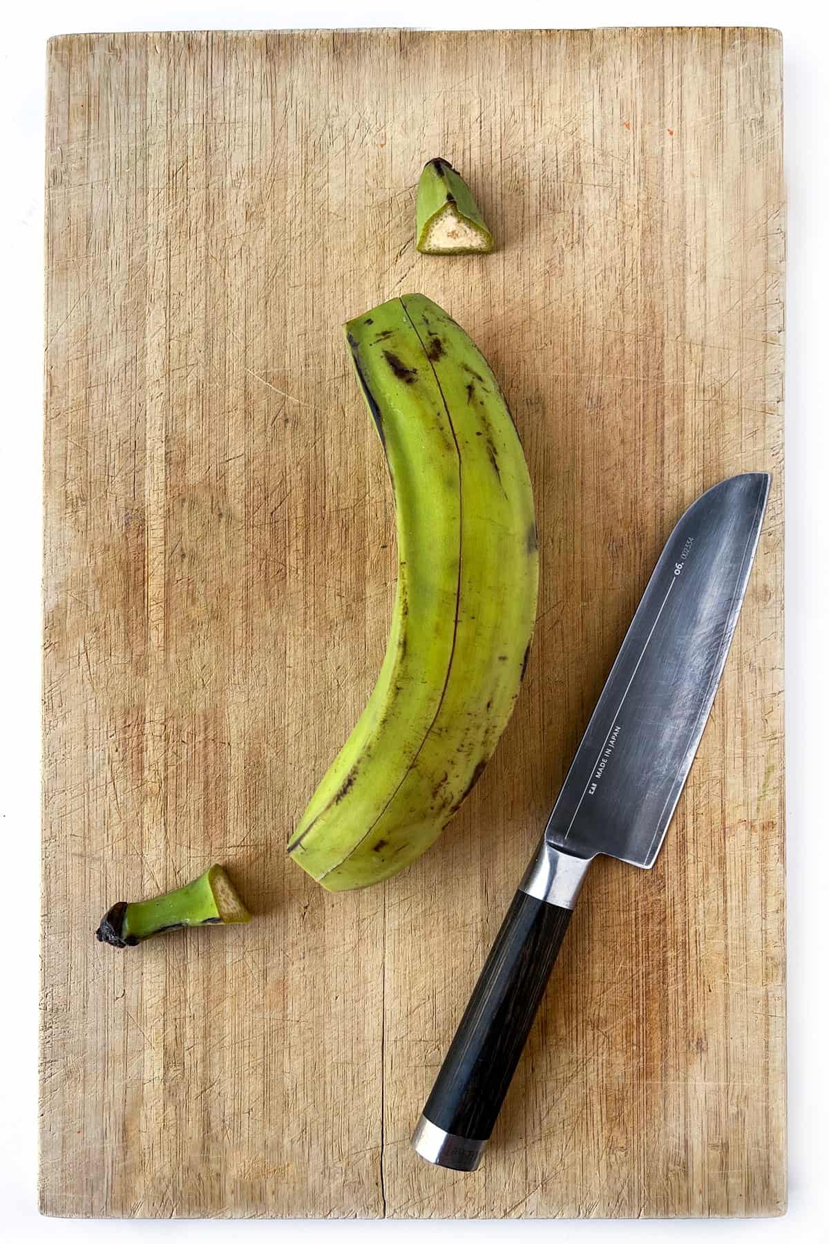 A green plantain with the ends snipped off and a slit cut into the skin from end to end, on a wooden cutting board with a knife