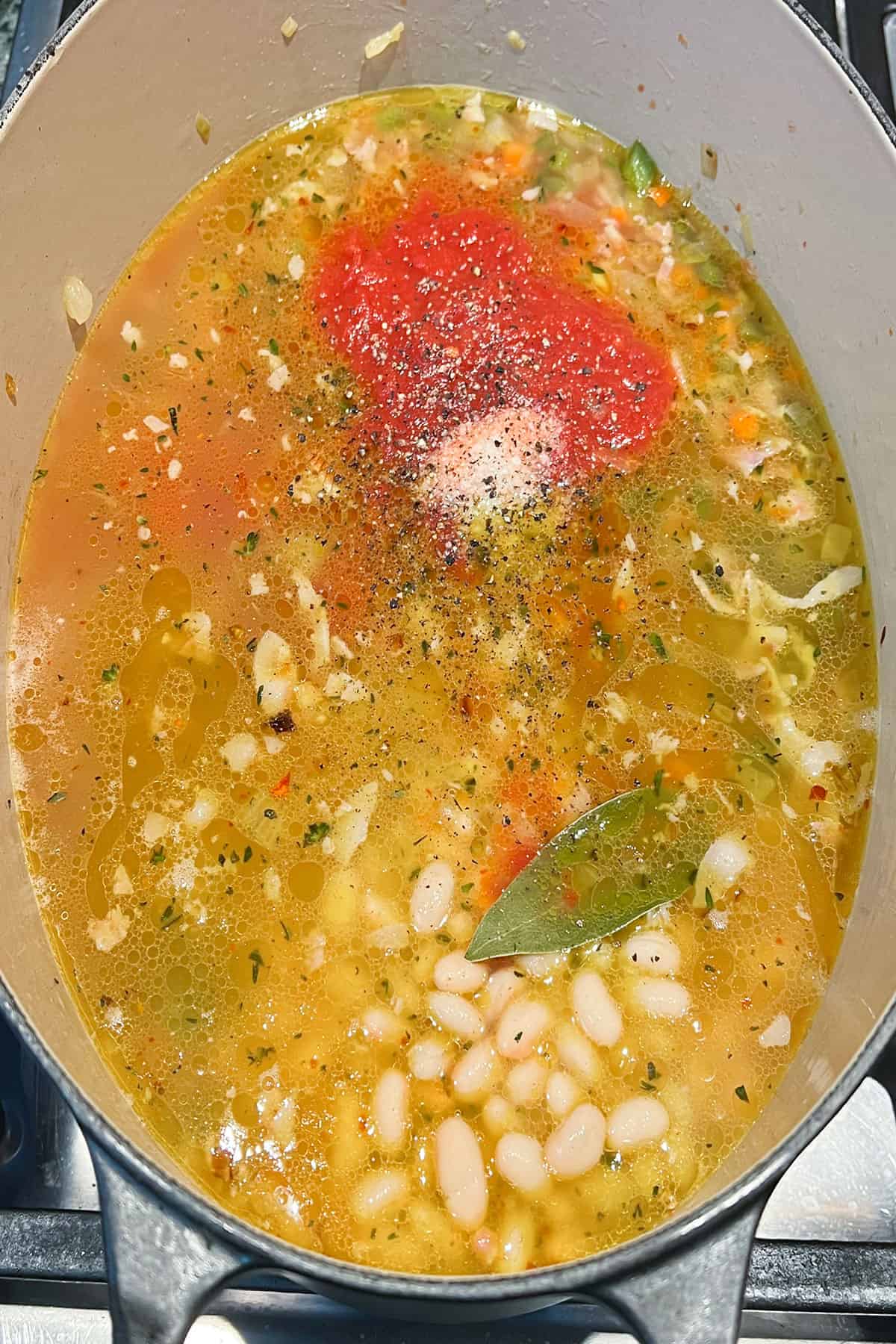 Dutch oven filled with broth, canned tomato puree, a bay leaf, white beans, and other ingredients for pasta and bean soup