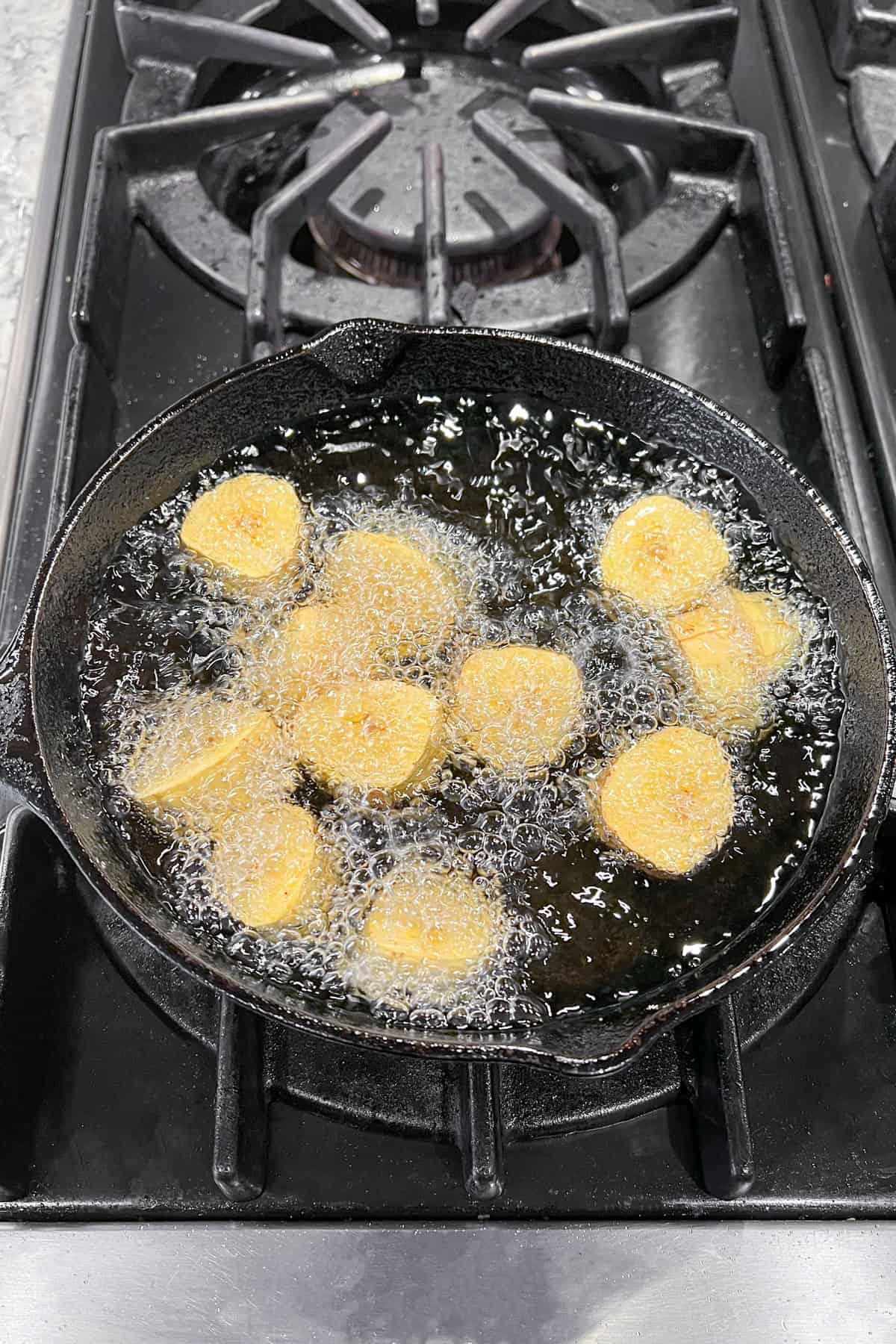 a dozen pieces of sliced green plantain frying in oil in a small cast iron skillet
