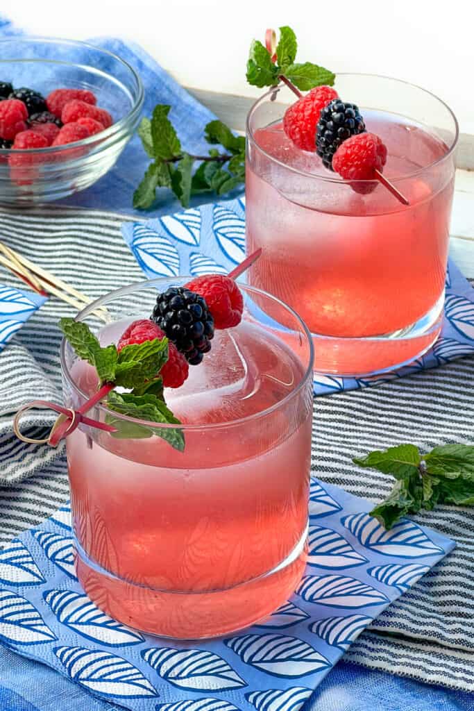 Two berry shrub drinks in glass tumblers with ice and garnished with toothpicks holding raspberries, blackberries and a mint sprigs