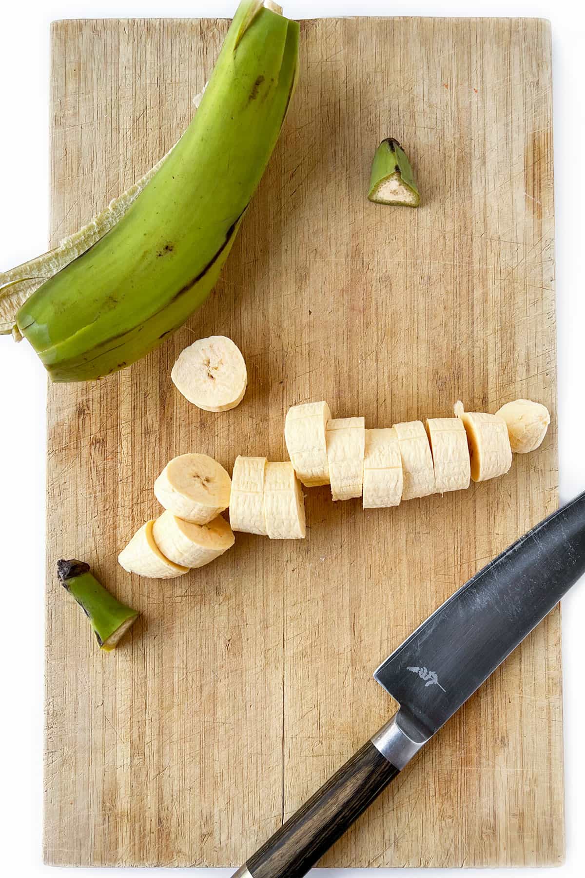 a sliced green plantain on a wooden cutting board, the skin and a knife off to the side