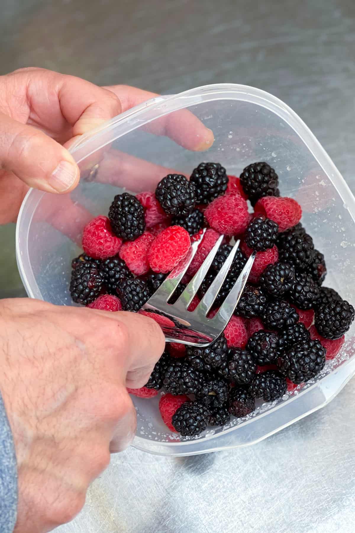 a large fork pressing down on fresh blackberries and raspberries that are in a plastic container