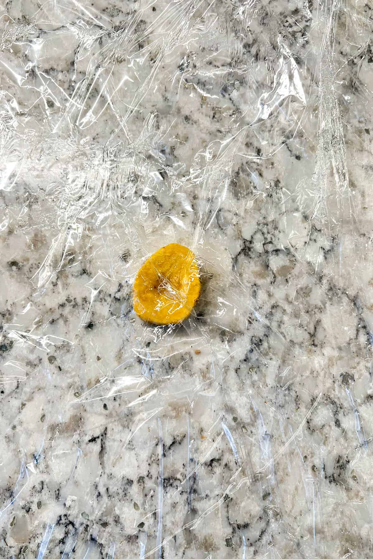 one small round disc of sliced plantain on a granite countertop green between two layers of plastic wrap