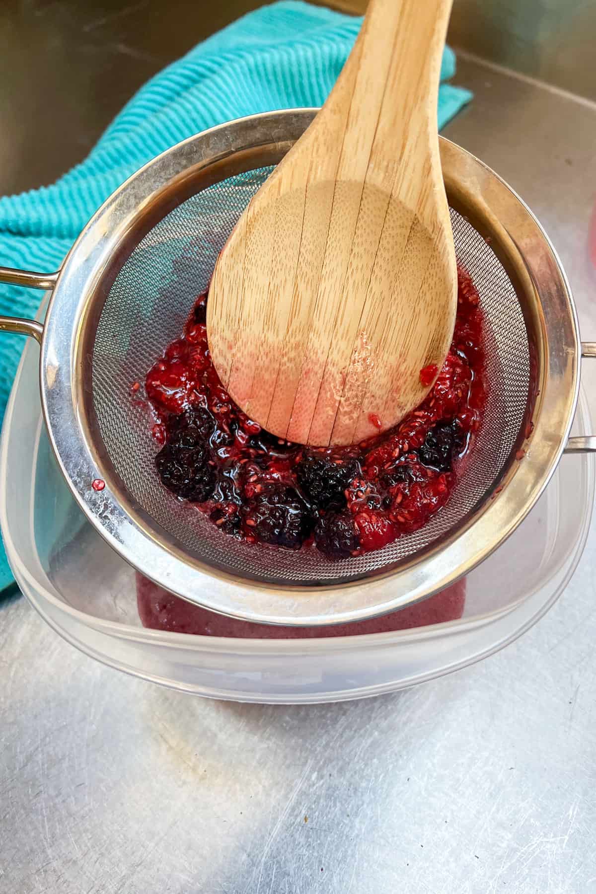 macerated berries in a mesh strainer being press on by a wooden spoon so the juices are pushed down into the container below