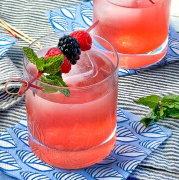 Berry shrub drink in a glass tumbler with a garnish on top of a toothpick with three berries and a mint sprig.