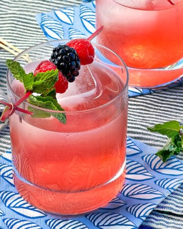 Berry shrub drink in a glass tumbler with a garnish on top of a toothpick with three berries and a mint sprig.