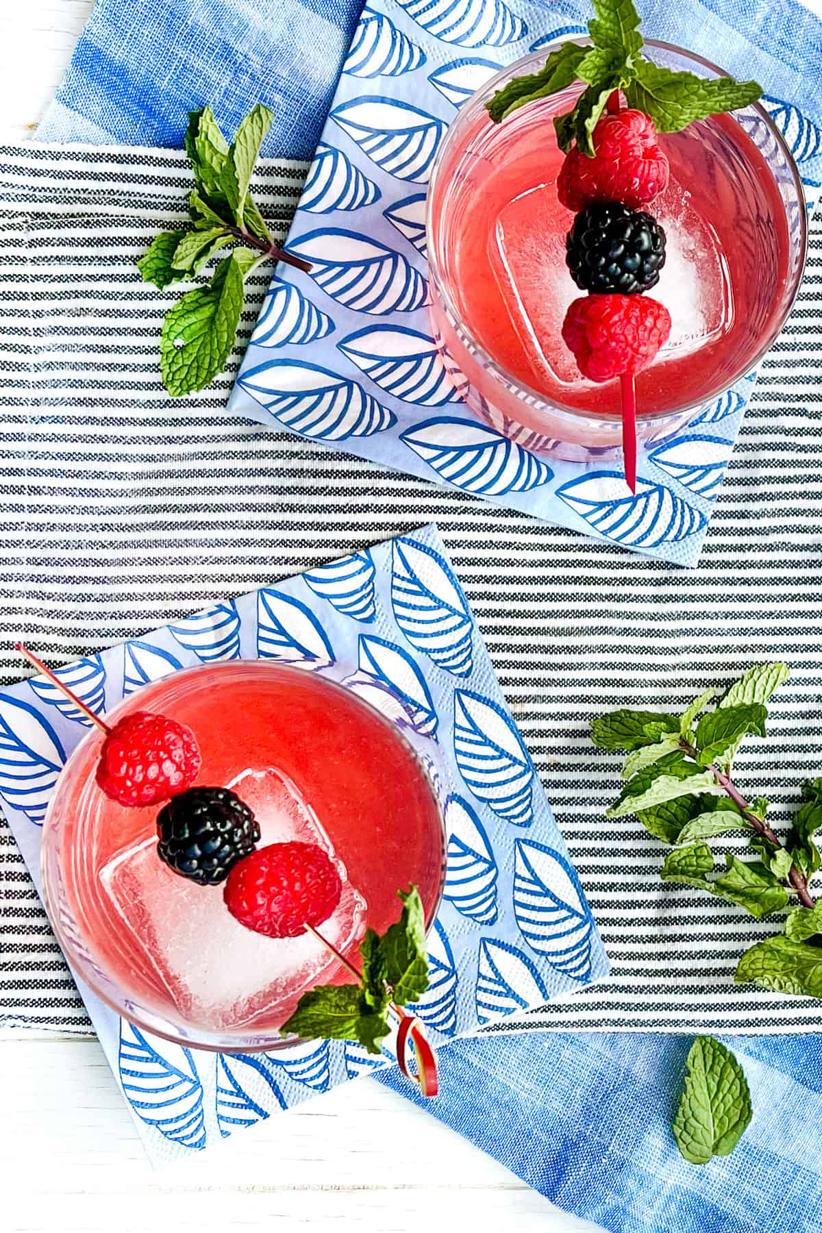 overhead looking down on two tumblers filled with ice and a berry shrub garnished with a toothpick holding three berries and a mint sprig
