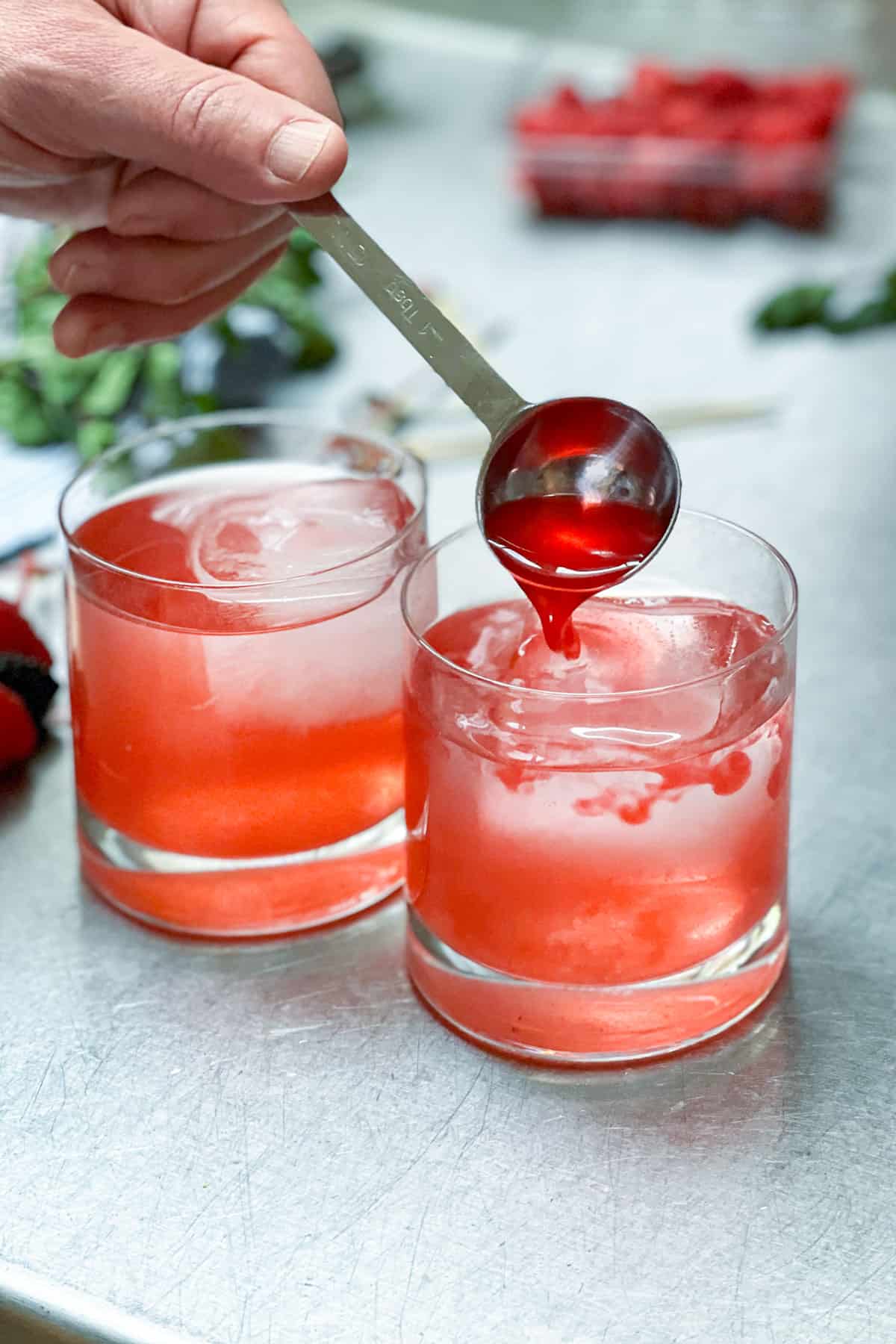 two glass tumblers filled with ice, a measuring spoon adding a tablespoon of shrub syrup to one of the glasses