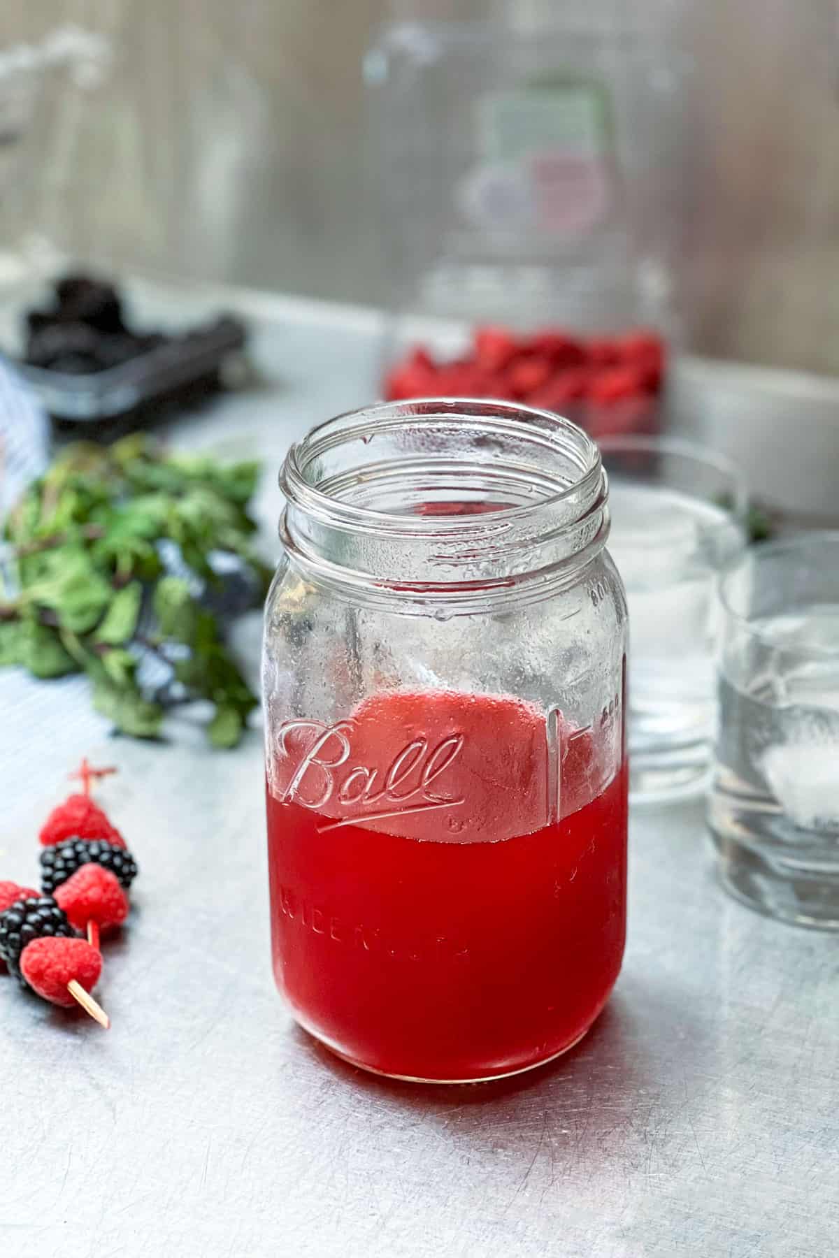 A glass mason jar half filled with berry shrub syrup, some ice filled glasses in the background and some herbs and  berries