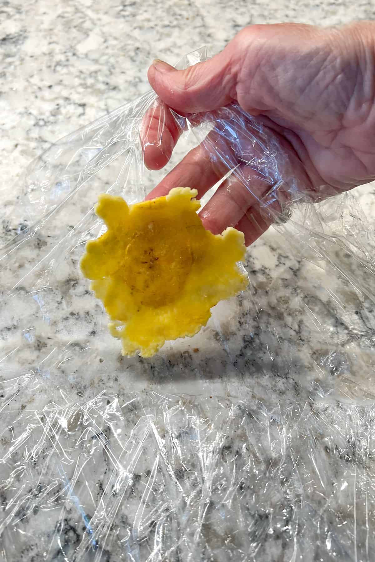 one flattened fried piece of green plantain being lifted off a piece of saran wrap