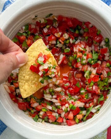 A corn tortilla chip being dipped into a bowl filled with finely chopped salsa fresca made from tomatoes, chilies, onion and cilantro.