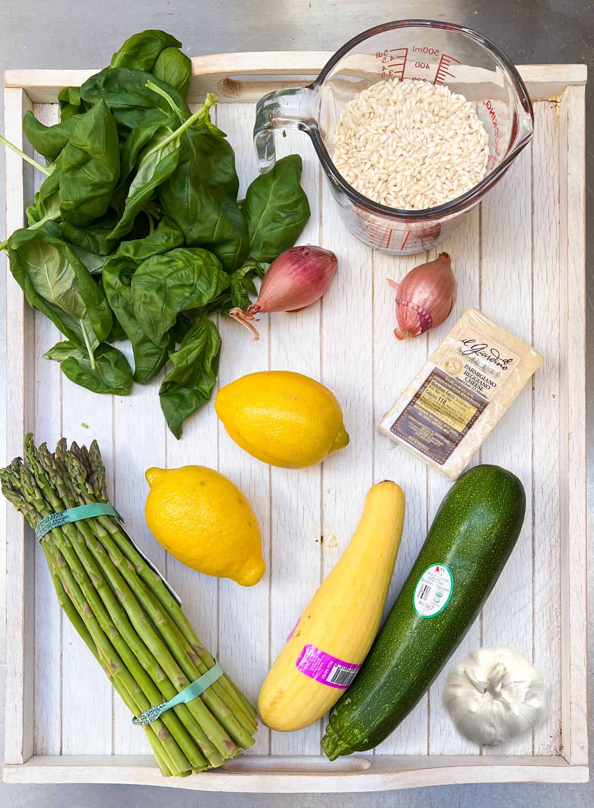 Risotto ingredients on a white wooden tray: two summer squash, a bunch of asparagus, 2 lemons, a bulb of garlic, a bunch of fresh basil, 2 shallots, a hunk of parmigiano-reggiano cheese, and a measuring cup filled with short grain rice