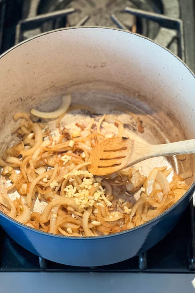 sliced onions and chopped garlic cooking in a dutch oven with a wooden spoon stirring