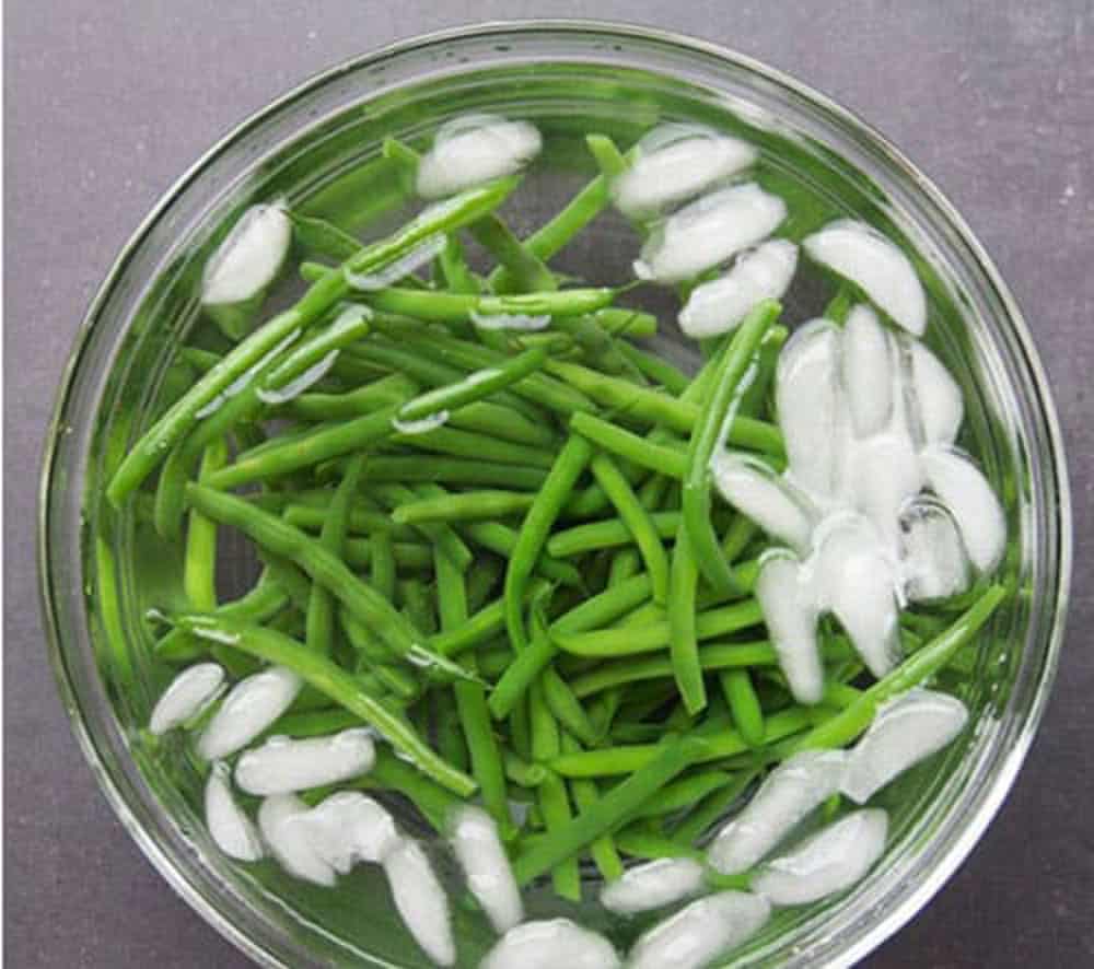 Blanched green beans in a glass bowl filled with ice water.