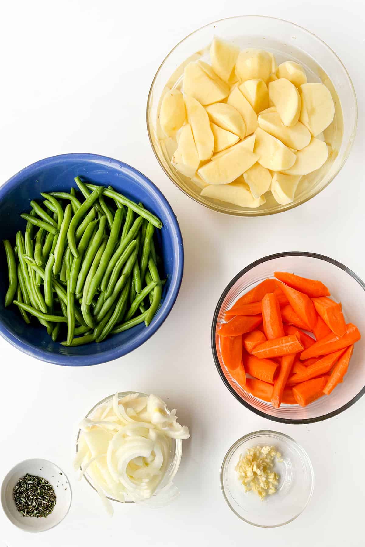 Glass bowl of potato wedges soaking in water, glass bowl of carrot wedges, small glass bowl of sliced onions, small glass bowl of chipped garlic, tiny bowl of finely chopped rosemary