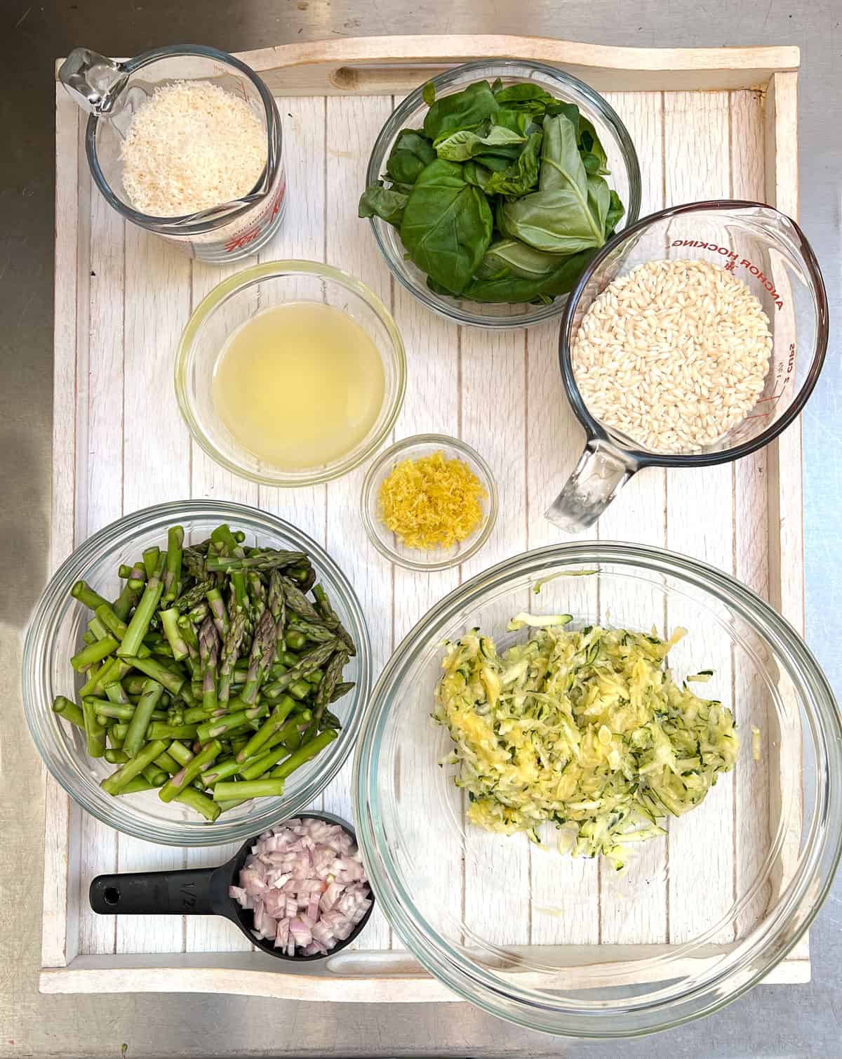 risotto ingredients, chopped, sliced and prepped on a white tray: chopped shallots, grated zucchini, sliced asparagus, leaves of basil in a bowl, small bowl of lemon juice, small bowl of lemon zest, cup of grated parmesan, cup of short grain rice
