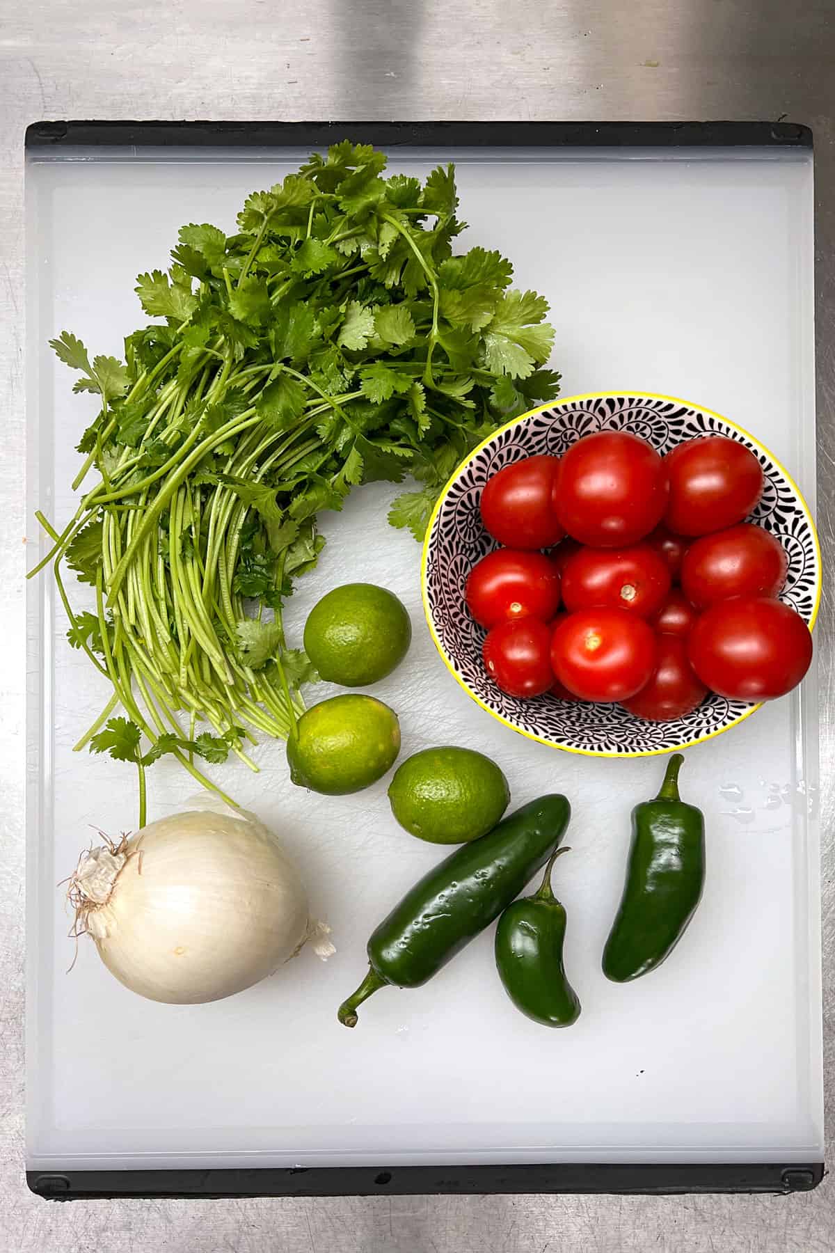 Salsa fresca recipe ingredients on a white cutting board: a bunch of cilantro, a bowl of ripe tomatoes, a white ohnion, 3 jalepeno peppers and 3 limes.
