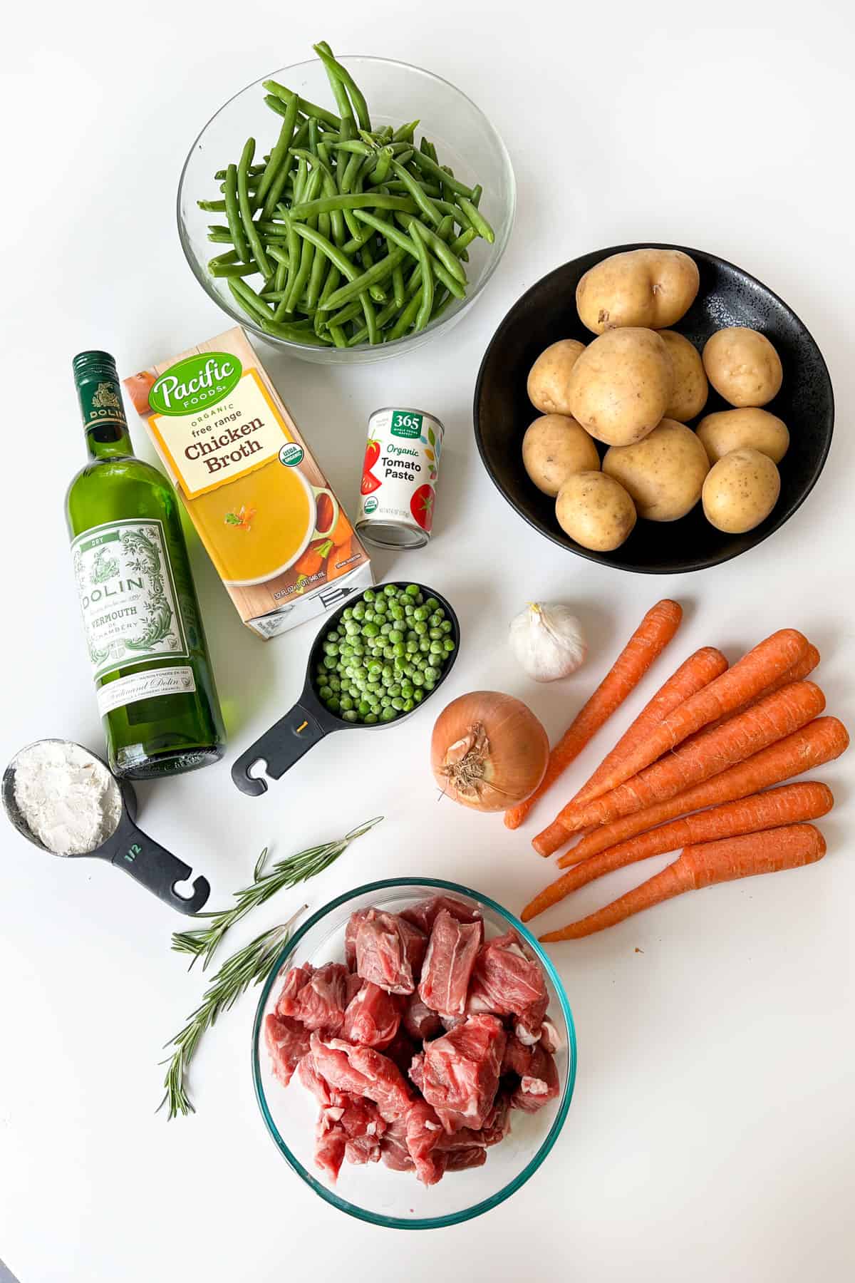 lamb stew ingredients on a white surface: bowl of green beans, bowl of yukon gold potatoes, bottle of dry vermouth, box of chicken broth, can of tomato paste, 8 carrots, a yellow onions, a bulb of garlic, a bowl of lamb stew meat, a cup of peas, a half cup of white flour, two sprigs of rosemary.