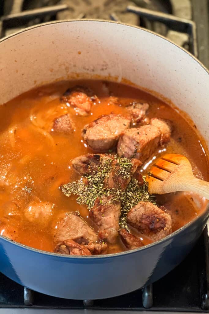 broth and chopped rosemary getting stirred into a pot of lamb stew with a wooden spoon.