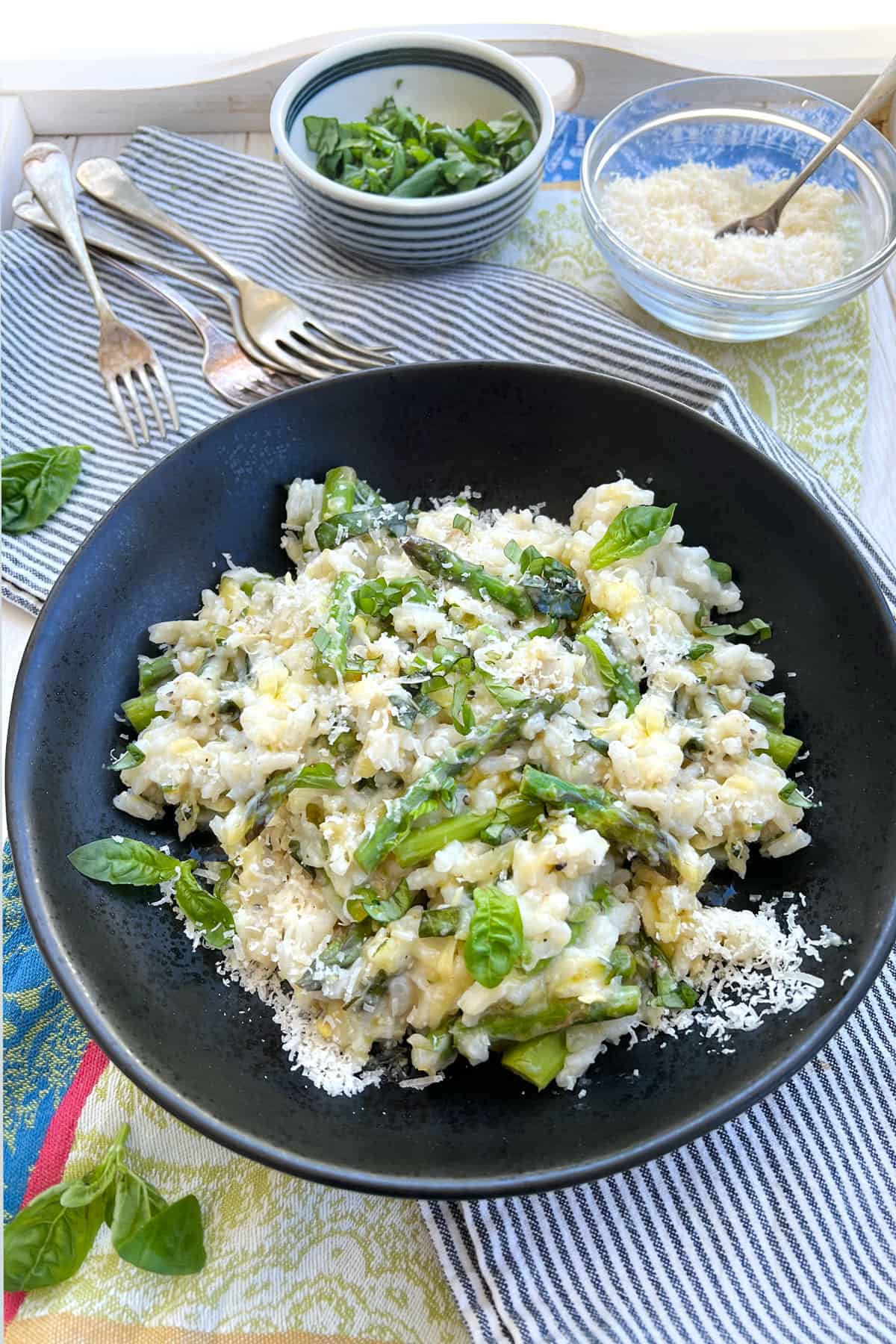 White wooden serving tray topped with a striped cloth napkin, toped with a plate of lemon asparagus risotto. Several forks on the tray and small bowls of parmesan and basil