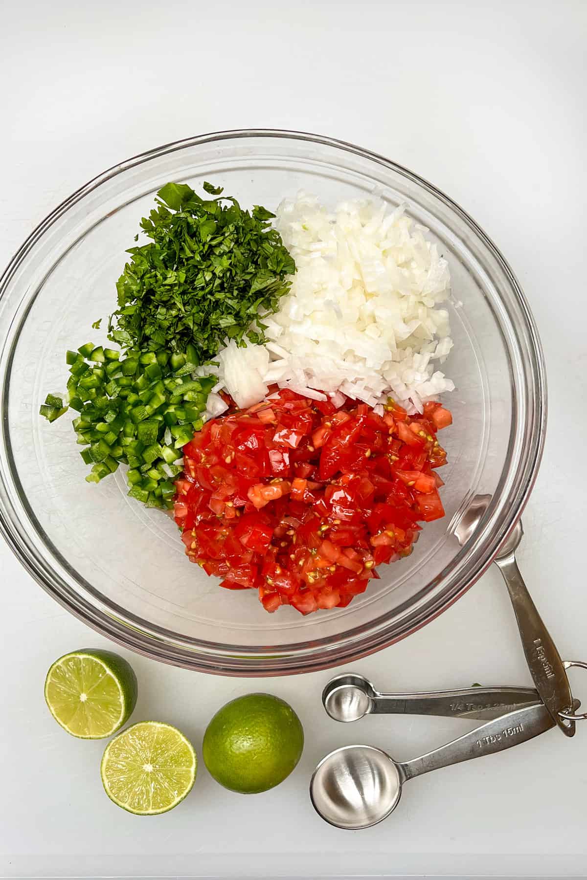 Salsa fresca recipe ingredients in separate piles in a glass bowl: diced onion, diced tomatoes, diced jalapenos and chopped cilantro.