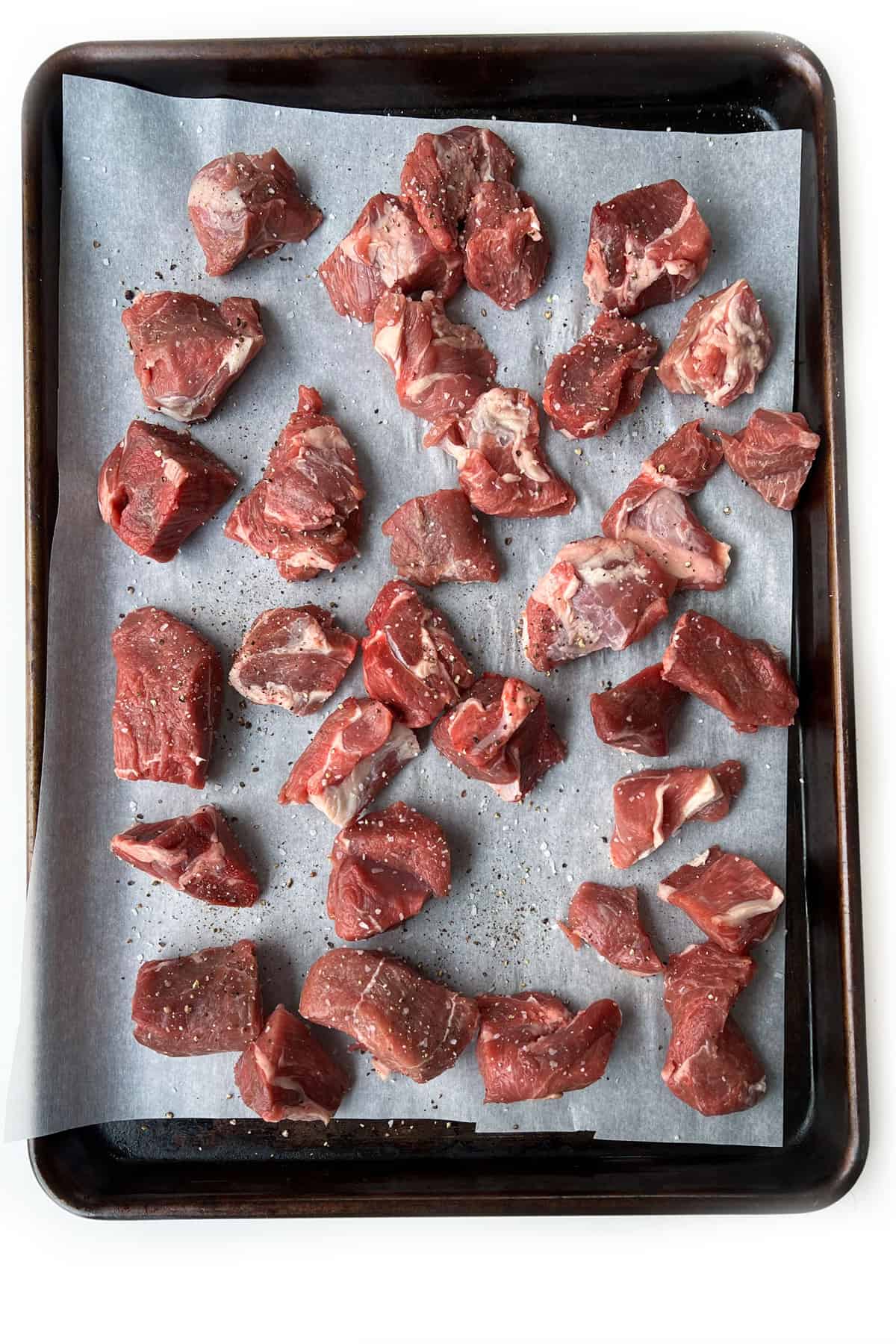 cubes of salted and peppered lamb stew meat spread out on a parchment lined rimmed baking sheet.