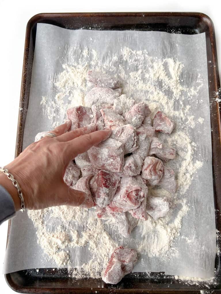 hand tossing floured chunks of lamb on a parchment lined rimmed baking sheet.