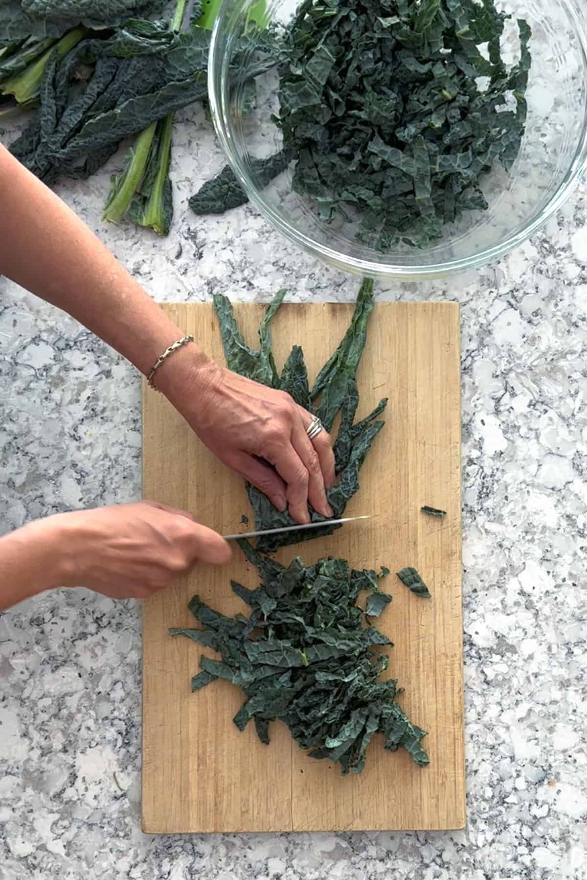 Hands shown slicing a stack of tuscan kale leaves, on a wooden cutting board, into thin slivers. A glass bowl of shredded tuscan kale sits next to the cutting board.