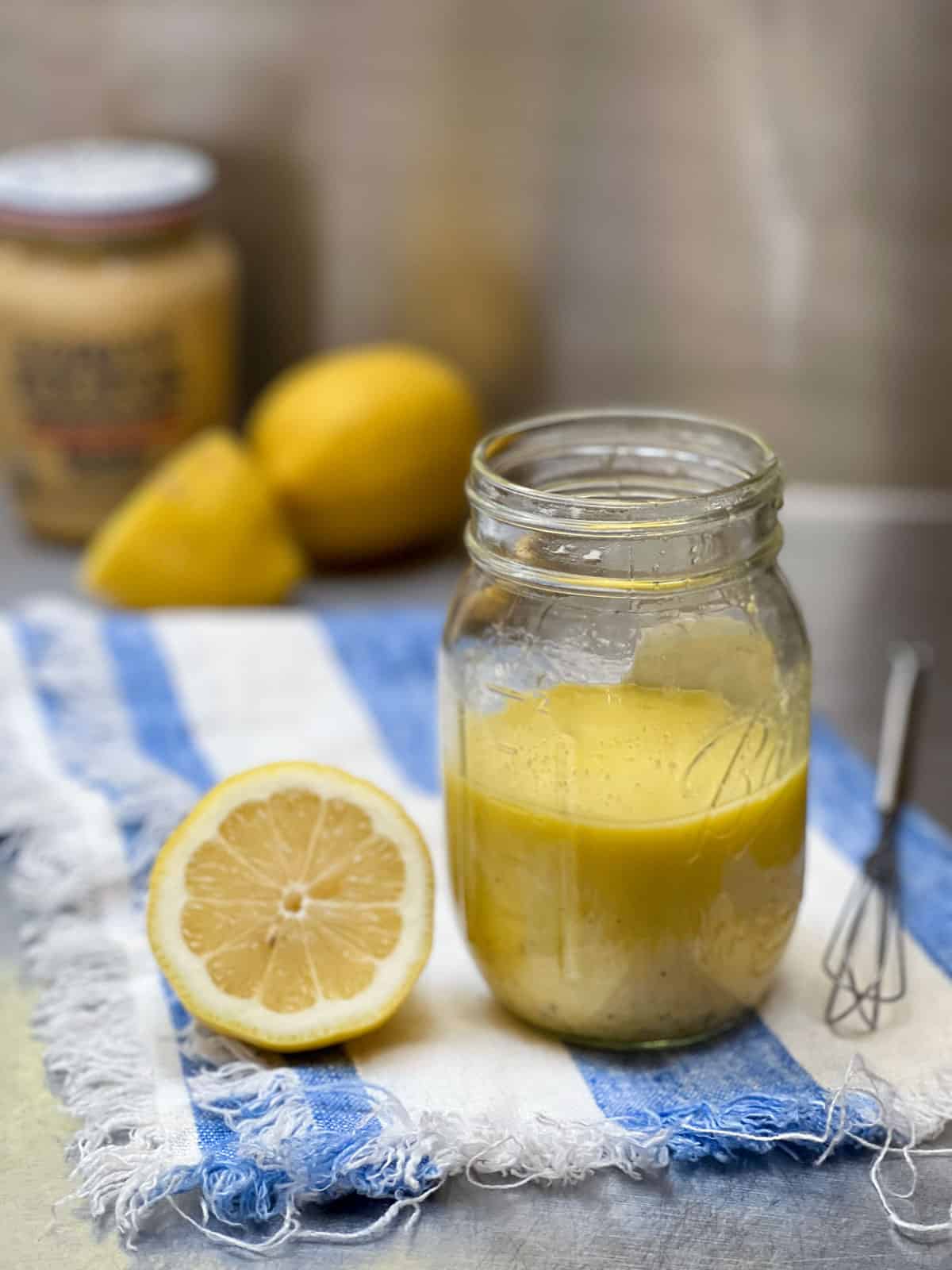 Lemon vinaigrette in a mason jar with a sliced half lemon next to it and two lemons in the background with a jar of dijon mustard.