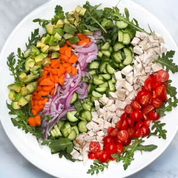 Rows of colorful chopped veggies and chicken in a serving bowl: cherry tomatoes, chicken, cucumbers, red onion, carrots and avocados.