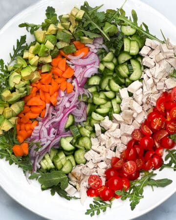 Rows of colorful chopped veggies and chicken in a serving bowl: cherry tomatoes, chicken, cucumbers, red onion, carrots and avocados.