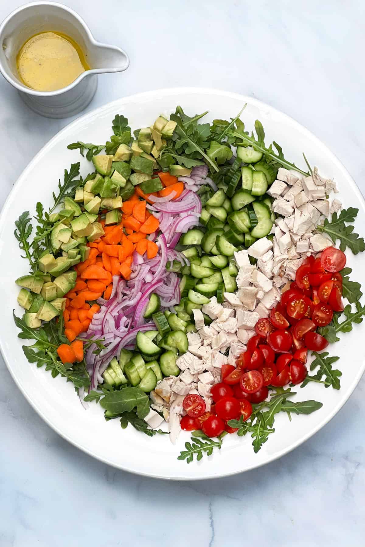 A rainbow of diced ingredients lined up in a large serving bowl, avocados, carrots, red onions, cucumbers, chicken, and cherry tomatoes. A pitcher of lemon dijon vinaigrette to the side.