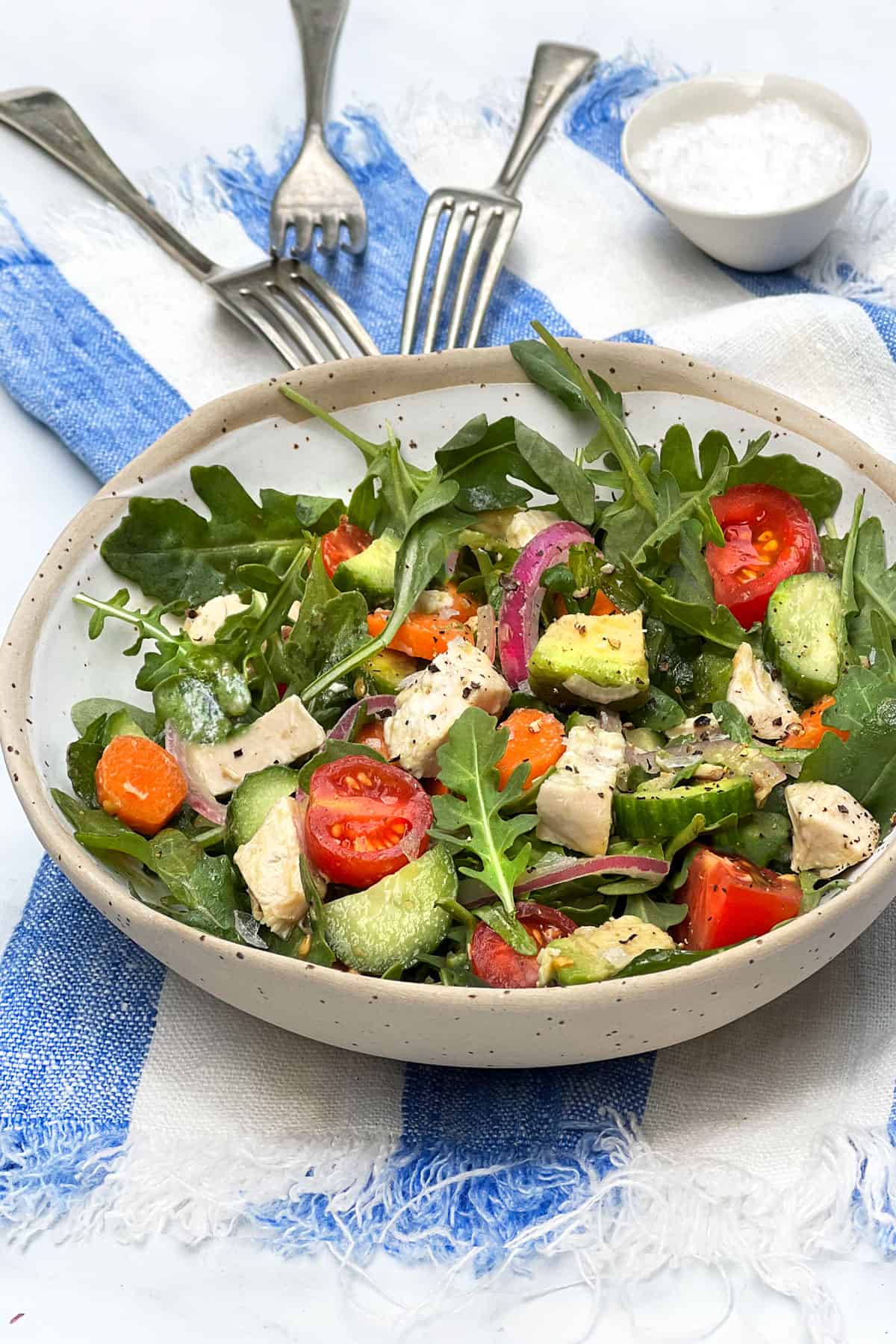 Bowl of chopped salad with chicken, cherry tomatoes, arugula, avocados, carrots and cucumbers, 3 forks and a small salt cellar behind the bowl