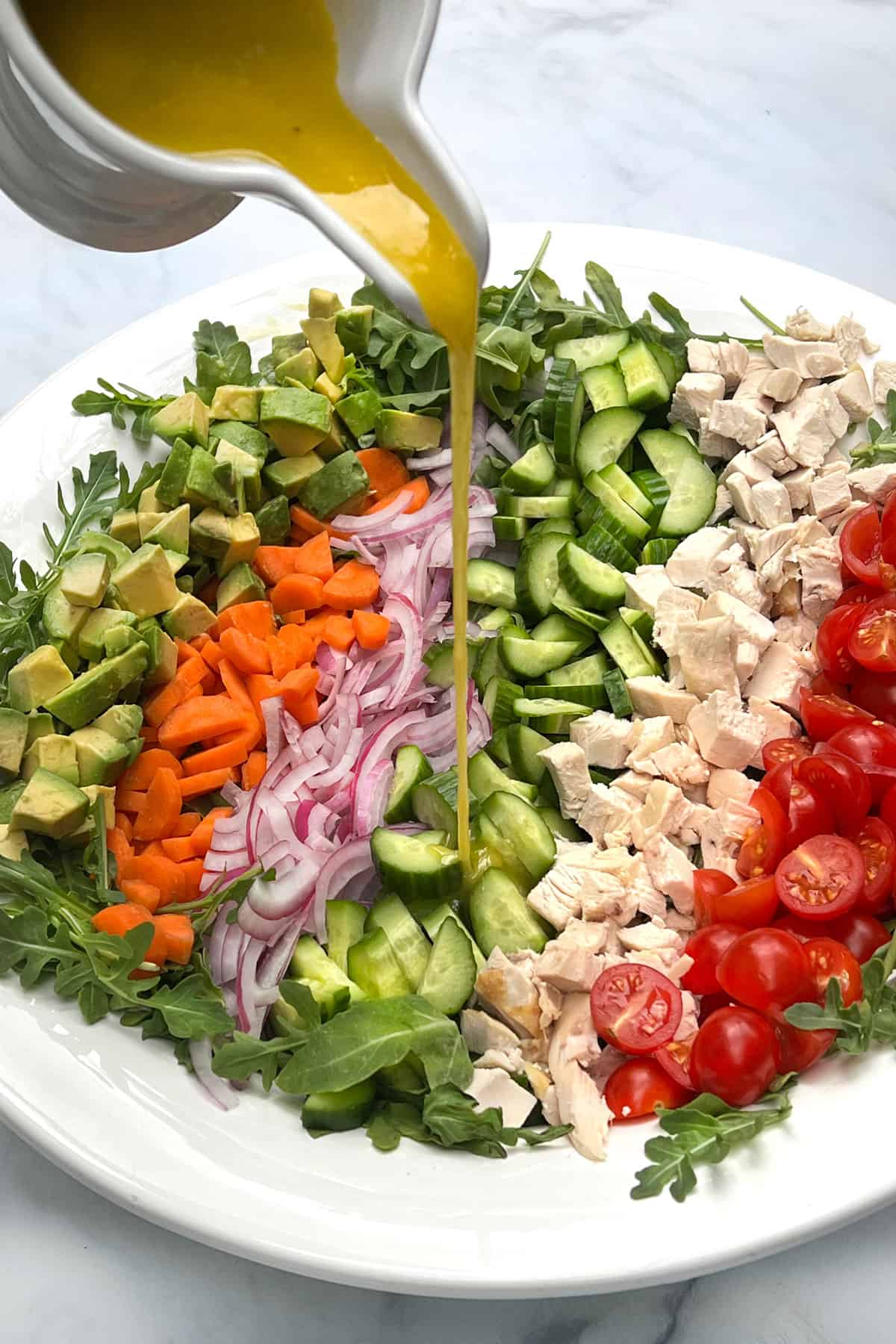 A rainbow of diced ingredients lined up in a large serving bowl, avocados, carrots, red onions, cucumbers, chicken, and cherry tomatoes. A stream of lemon dijon vinaigrette is being poured on top.