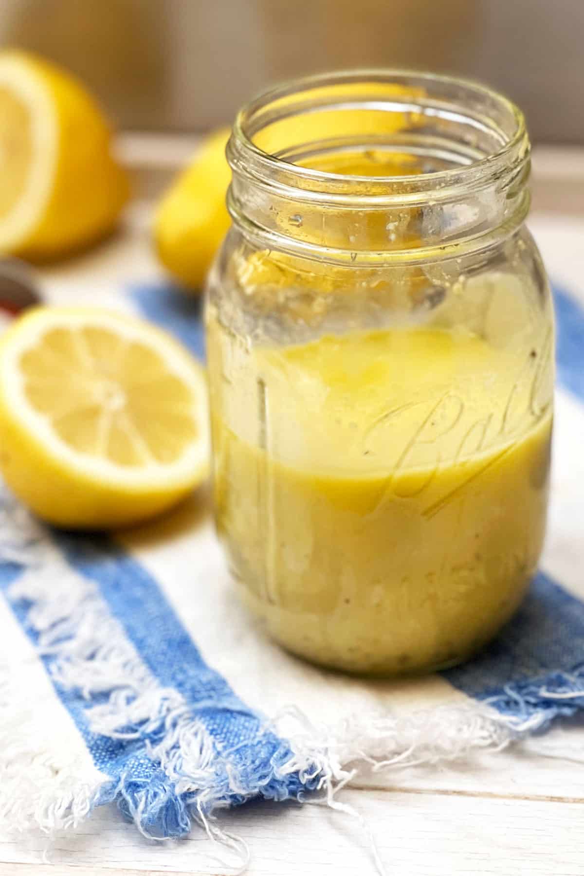 A mason jar on a striped cloth napkin, filled halfway with lemon vinaigrette, half a sliced lemon sitting next to it and a few lemons in the background, small wire whisk to the side
