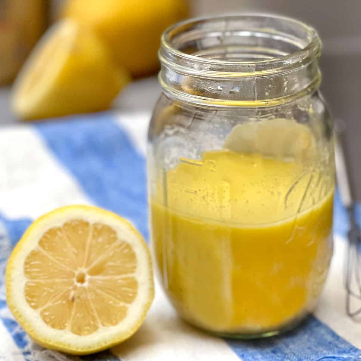 A mason jar on a striped cloth napkin, filled halfway with lemon vinaigrette, half a sliced lemon sitting next to it and a few lemons in the background, small wire whisk to the side