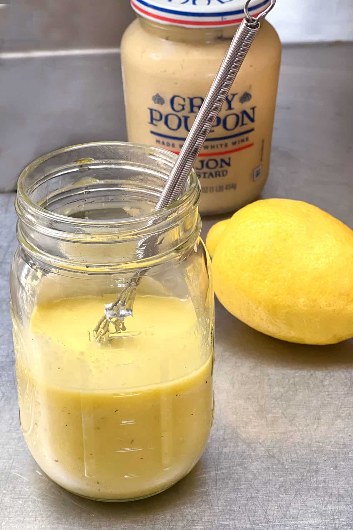 lemon vinaigrette in a mason jar with a small wire whisk in the dressing, a lemon on the side and a jar of dijon mustard in the background