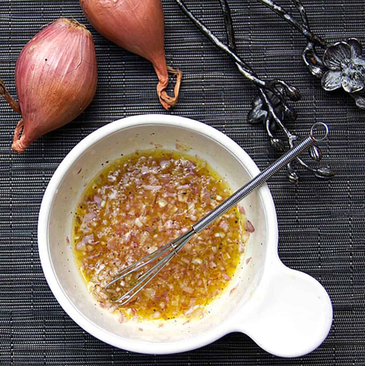 A small bowl filled with lemon honey shallot dressing, a small whisk resting in the bowl and two shallows to the side of the bowl