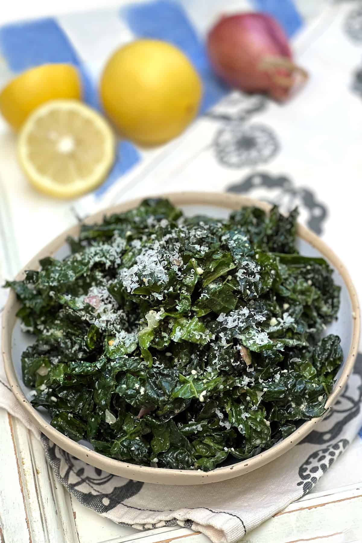 bowl of kale salad sprinkled with parmesan, a cut lemon and a shallot in the background